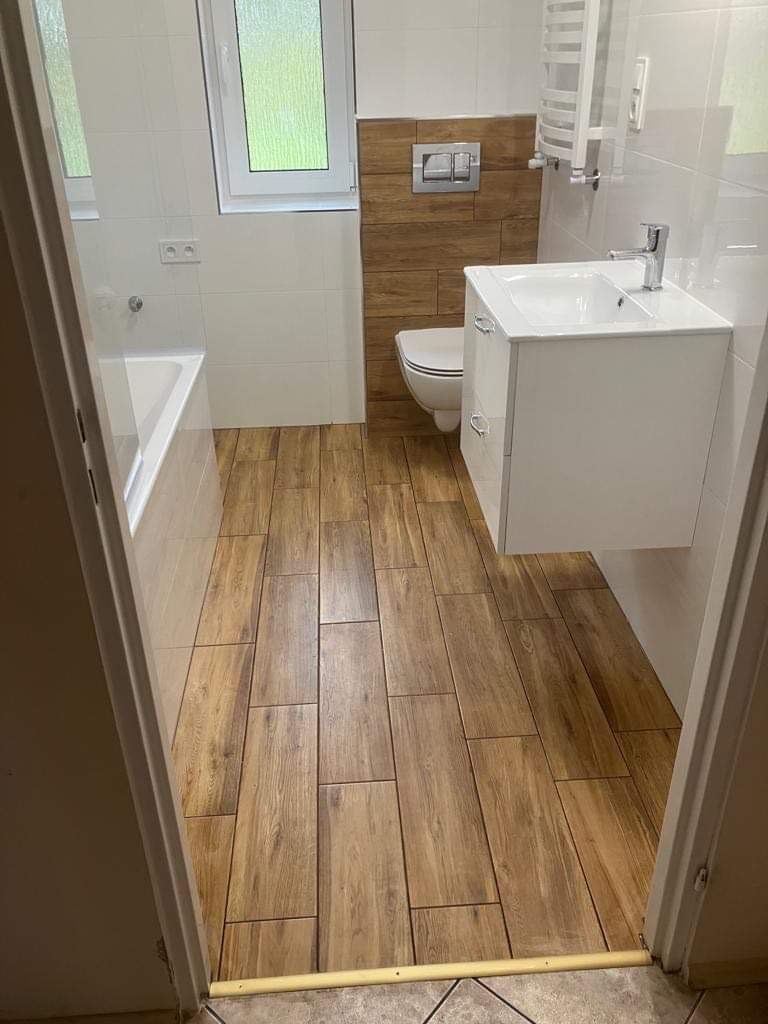 Jasna łazienka z drewnopodobną podłogą i białą armaturą. Widok zza progu, na ścianach białe płytki, akcent z drewnianych paneli za toaletą i nad nią.