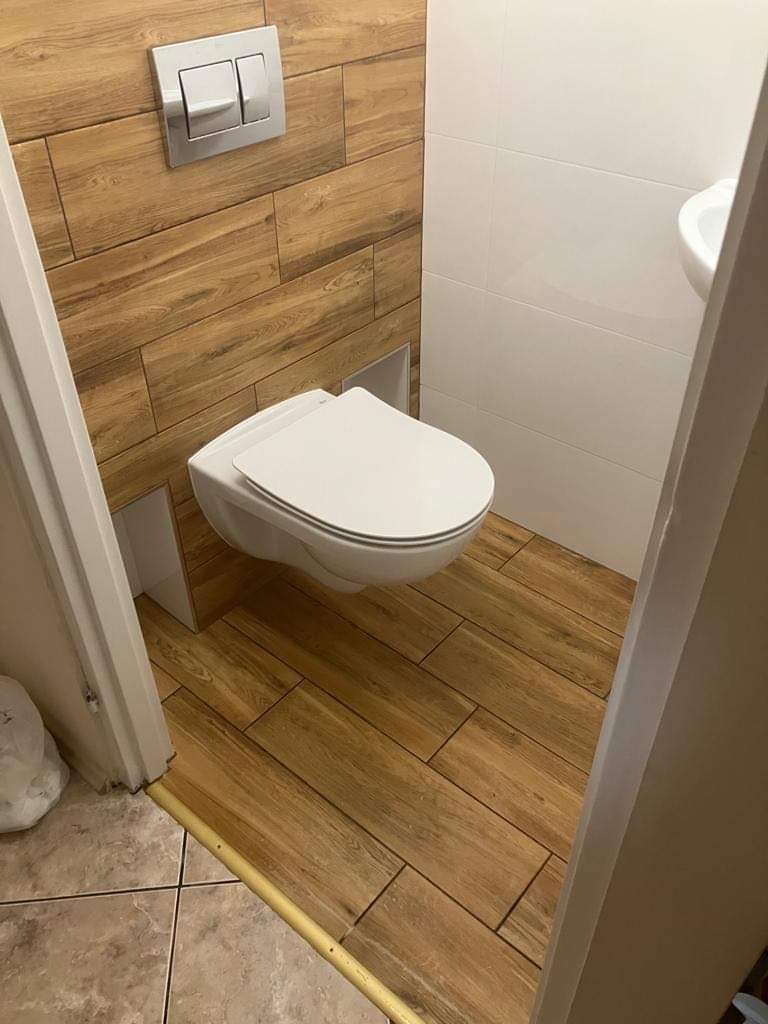 Nowoczesna łazienka z podwieszaną toaletą, drewnopodobnymi płytkami na ścianie i podłodze, oraz białymi płytkami na pozostałej części ścian.