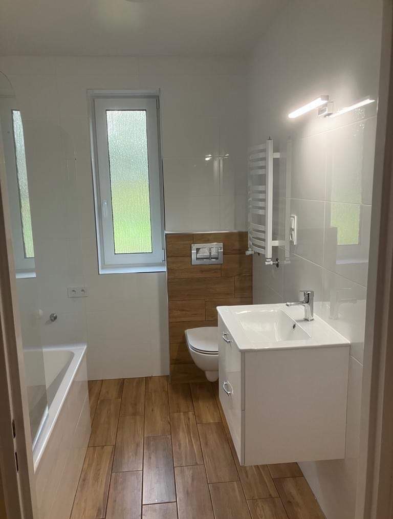 Nowoczesna łazienka z drewnopodobnymi płytkami, białą armaturą i grzejnikiem drabinkowym. Okno z widokiem na zieleń.