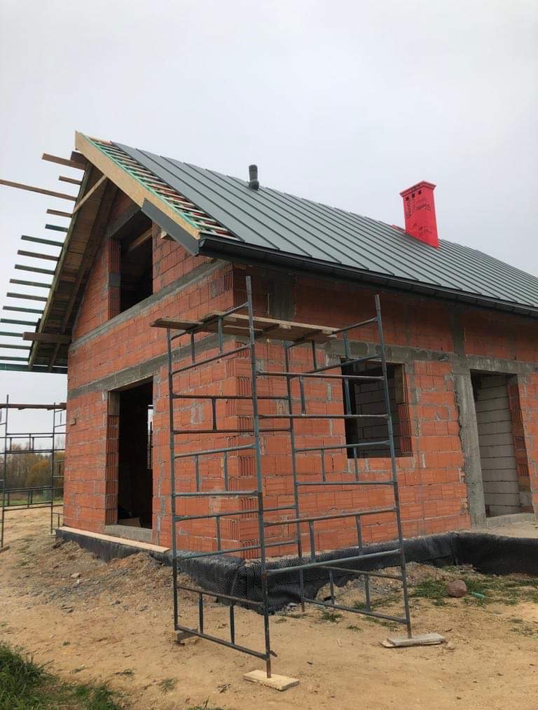 Surowy, dwukondygnacyjny dom z czerwonej cegły w trakcie budowy. Na dachu szara blacha, czerwony komin. Widoczne rusztowania i elementy konstrukcji dachu.