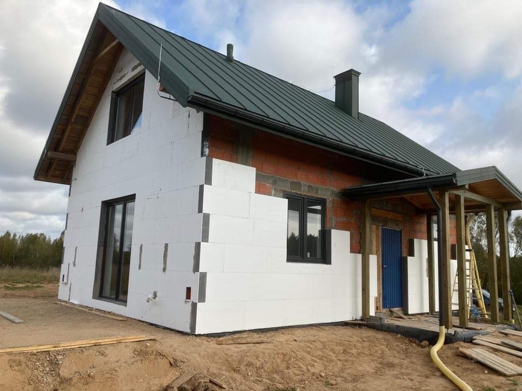 Dom w trakcie ocieplania styropianem, widoczne fragmenty ściany z cegły. Dach pokryty blachą w kolorze ciemnej zieleni. Okna czarne.