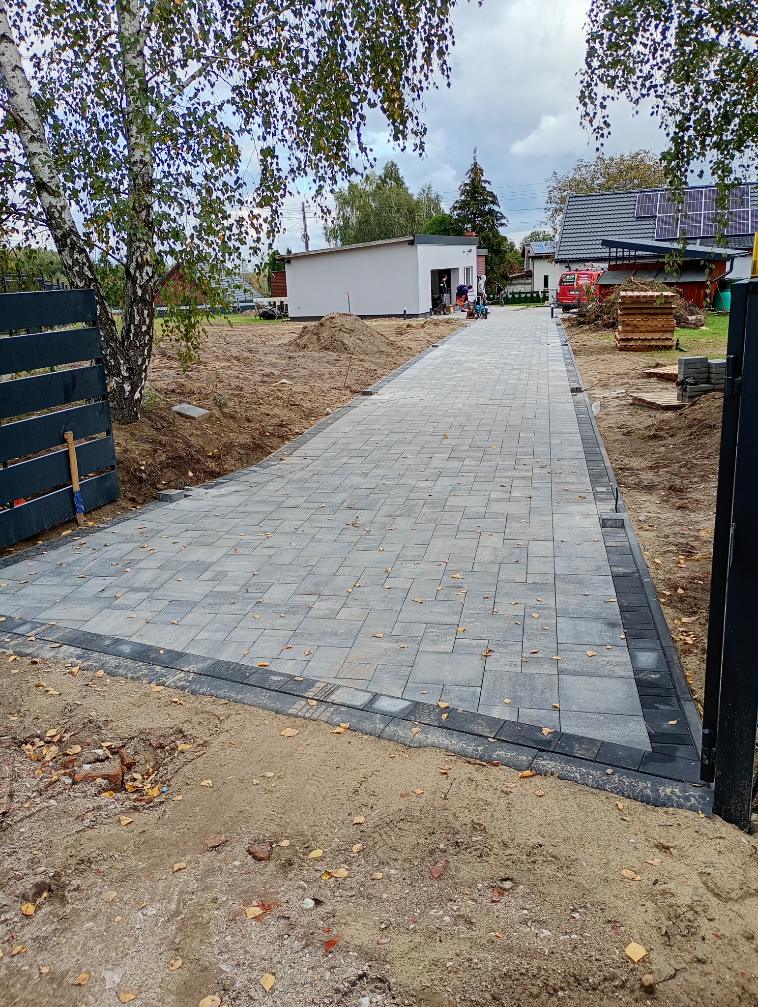 Ułożona ścieżka z kostki brukowej w odcieniach szarości, obramowana ciemniejszą kostką. W tle dom z panelami słonecznymi i ekipa brukarska.