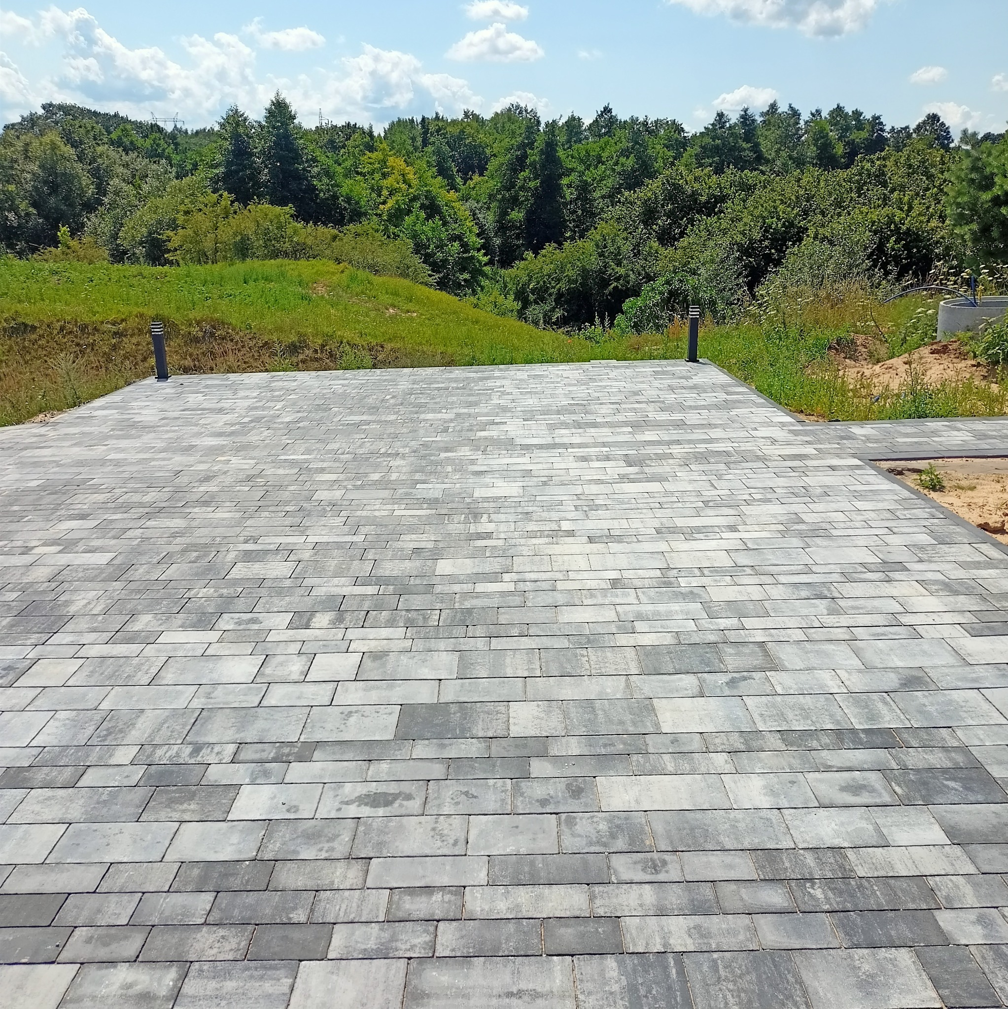 Ułożona szara kostka brukowa na posesji, z widokiem na zieleń i błękitne niebo. Widoczne dwie lampy oświetleniowe. Perspektywa z dołu, ukazująca wzór kostki.