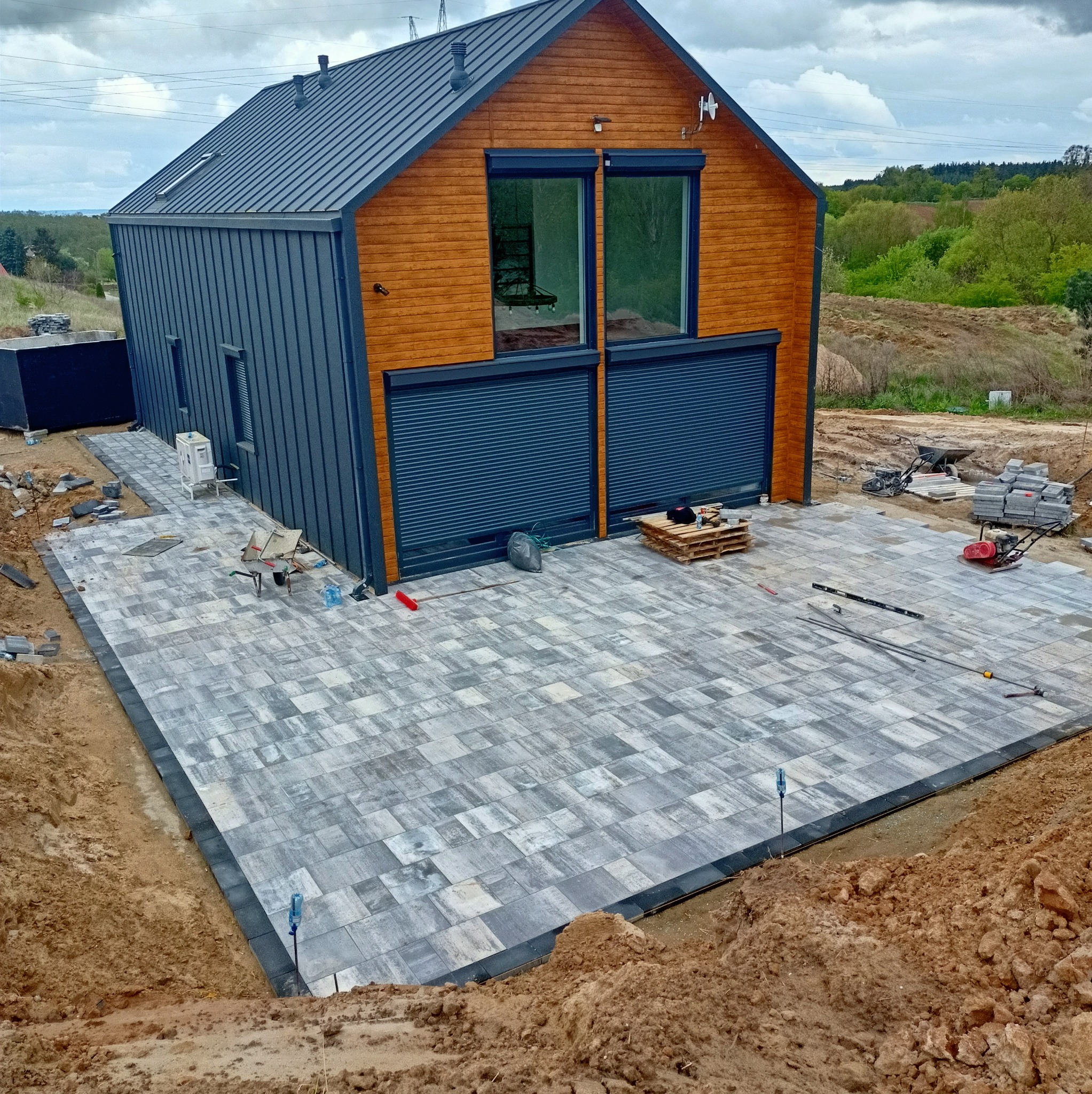 Nowoczesny dom z szarą kostką brukową na posesji. Widoczne narzędzia i materiały budowlane. Częściowo ułożona nawierzchnia z kostki w odcieniach szarości i bieli.