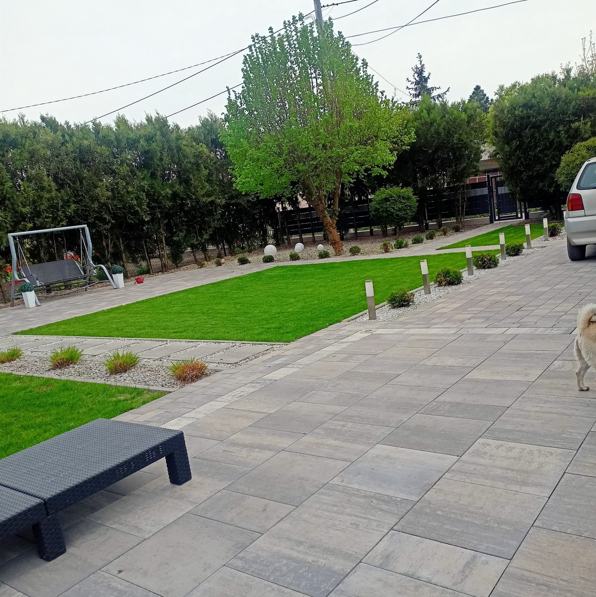 Szara kostka brukowa na podwórku z trawnikiem, oświetleniem i meblami ogrodowymi. Widoczny fragment samochodu i pies. Całość w spokojnej, stonowanej kolorystyce.