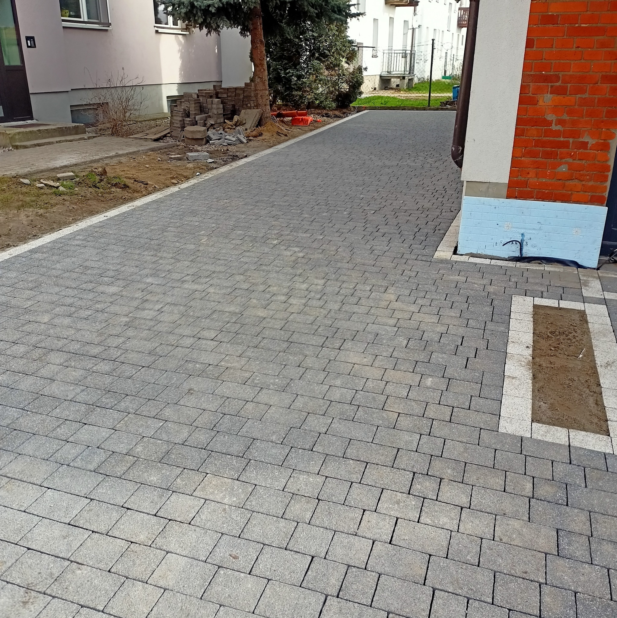 Ułożona szara kostka brukowa z białą obwódką przy budynku z czerwonej cegły. Na poboczu sterta kostki do dalszego układania. Widok z perspektywy.