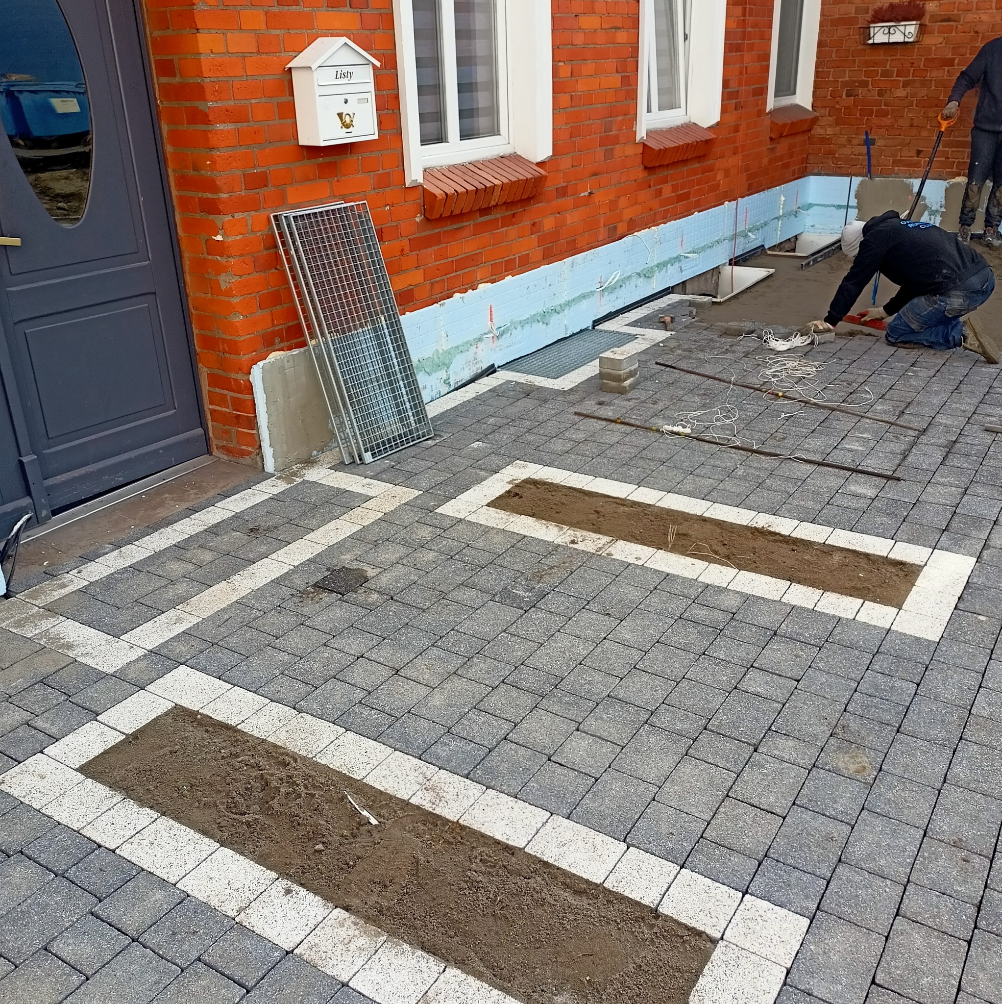 Montaż kostki brukowej w ogrodzie z elementami drenażu liniowego, widoczny etap prac z przygotowaniem podłoża. Ciekawy wzór ułożenia kostki w dwóch kolorach.