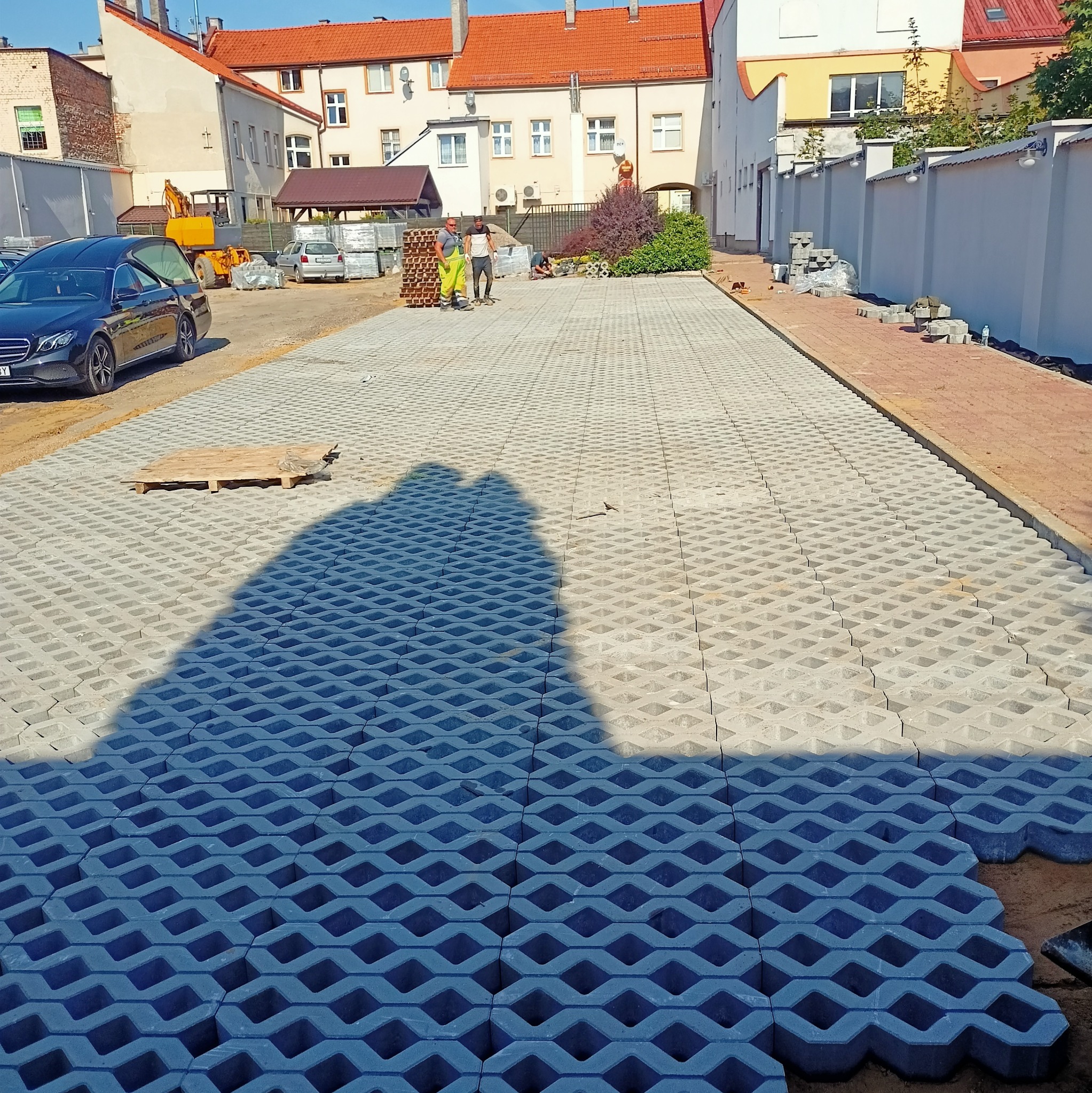 Nierównomiernie ułożona, szara ażurowa kostka brukowa na placu budowy, z paletą w tle i ekipą brukarzy. Częściowo widoczny czarny samochód.