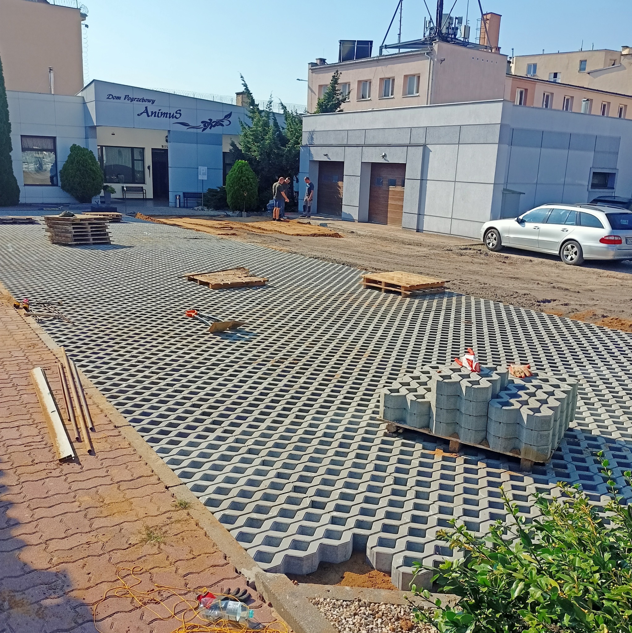 Montaż ażurowej kostki brukowej na posesji z budynkiem Domu Pogrzebowego w tle. Widoczne palety z kostką i narzędzia. Częściowo ułożona nawierzchnia.