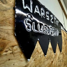 Mateusz Profus - Czarny, metalowy szyld z napisem 'Warsztat Ślusarski', przymocowany do ściany z płyty OSB. Szyld ma dekoracyjne wycięcia w dolnej części.