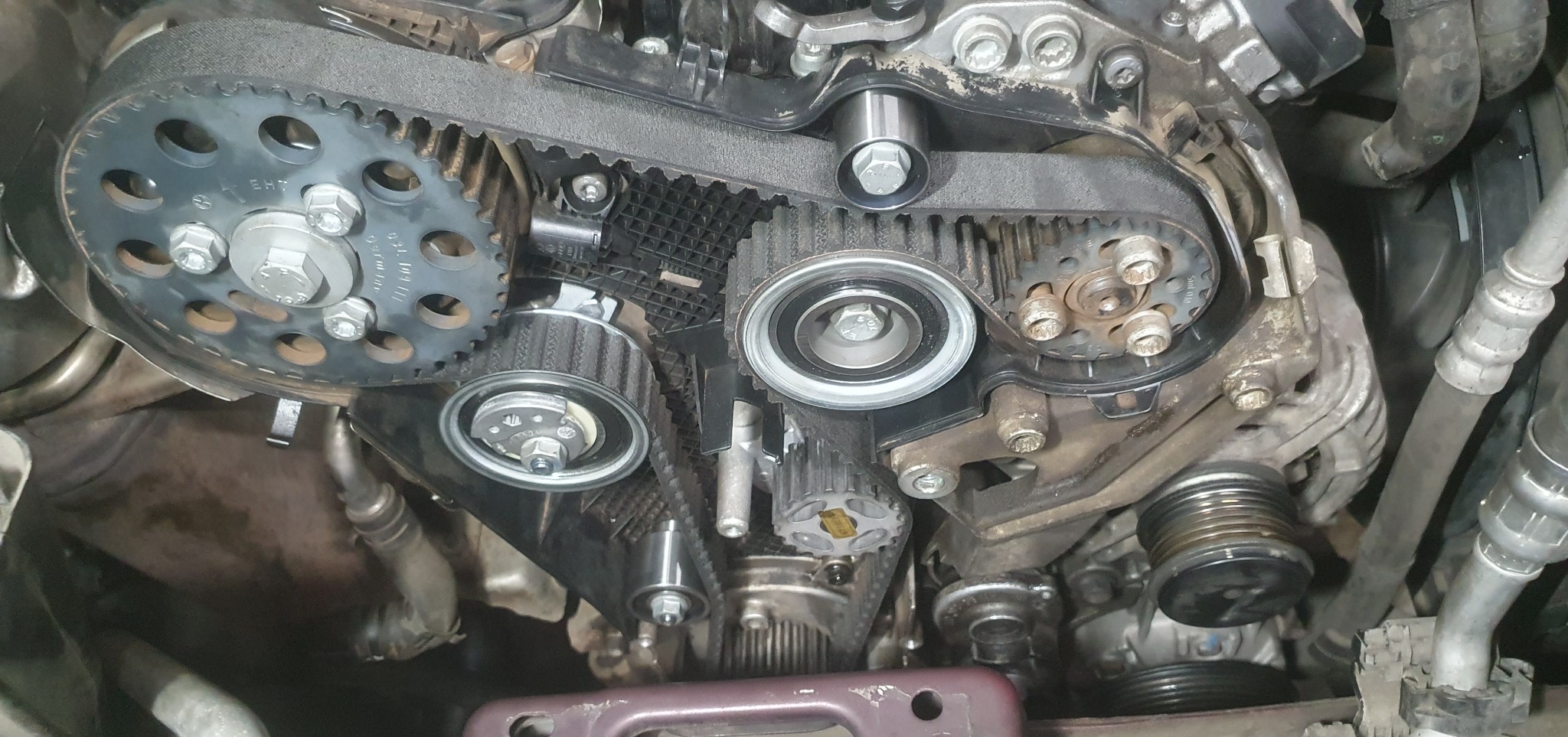 Widok z bliska na silnik samochodowy z widocznym paskiem rozrządu, kołami zębatymi i napinaczami. Elementy są brudne, co sugeruje użycie i potrzebę konserwacji.