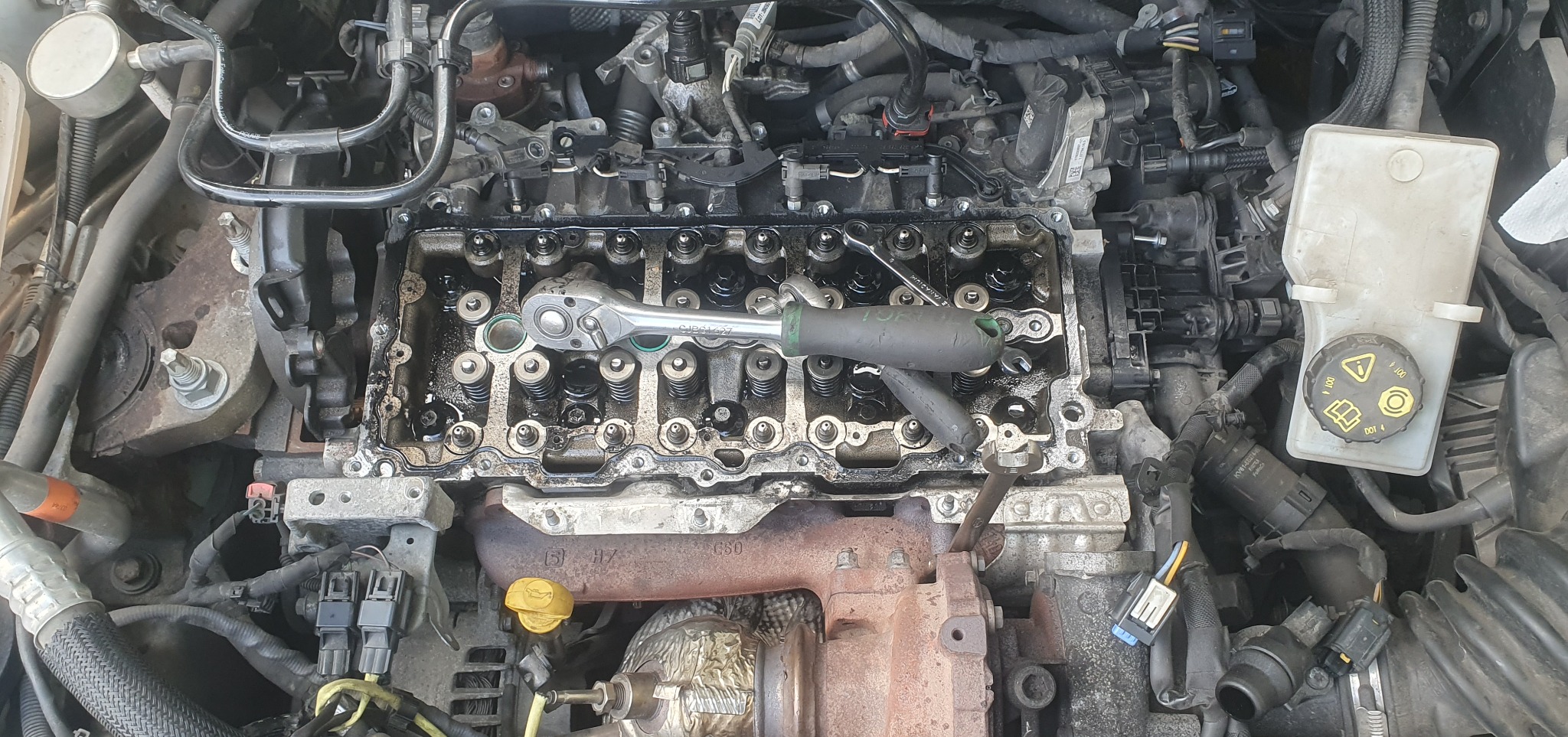 Otwarty silnik samochodowy z widocznymi sprężynami zaworowymi i nasadką klucza, sugerujący naprawę lub wymianę uszczelki pod głowicą. Widoczne przewody i elementy osprzętu.