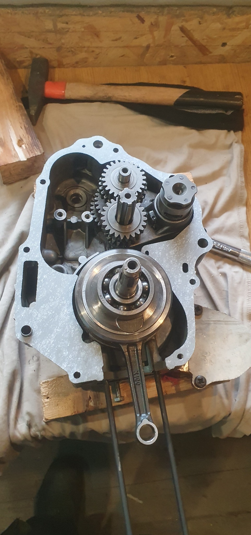 Silnik motocykla w trakcie naprawy: widoczny wał korbowy, skrzynia biegów. Młotek w tle sugeruje warsztatowy charakter. Detal mechaniczny, naprawa, serwis.