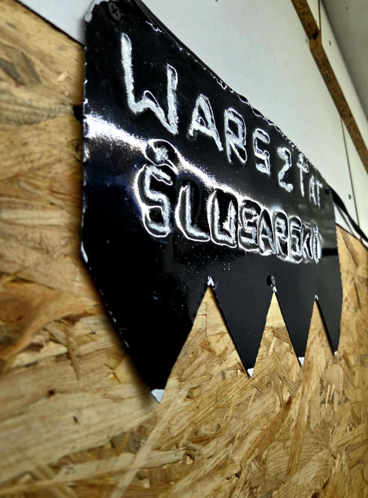 Czarny, metalowy szyld z napisem 'Warsztat Ślusarski', przymocowany do ściany z płyty OSB. Szyld ma dekoracyjne wycięcia w dolnej części.