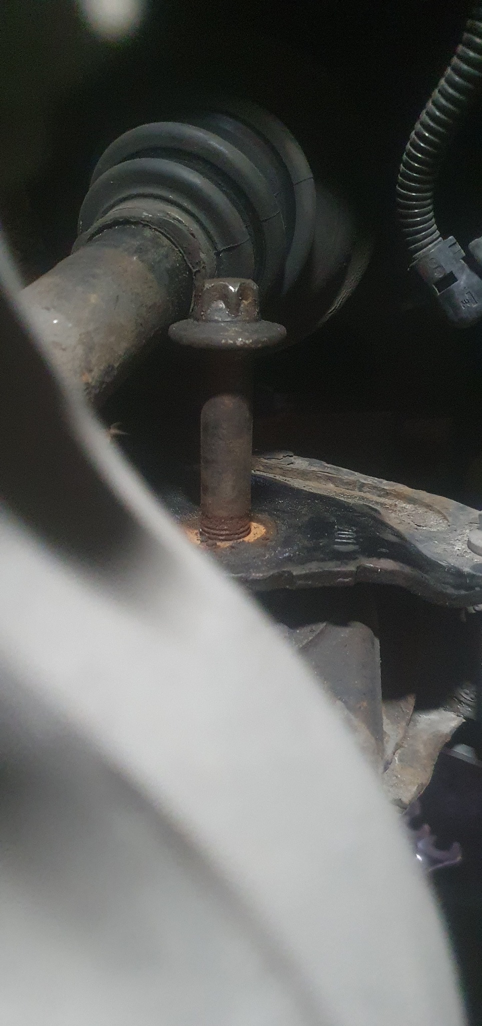 Rdzewiejąca śruba z nakrętką w zawieszeniu pojazdu, widoczne elementy gumowe i przewody. Niewyraźne ujęcie.