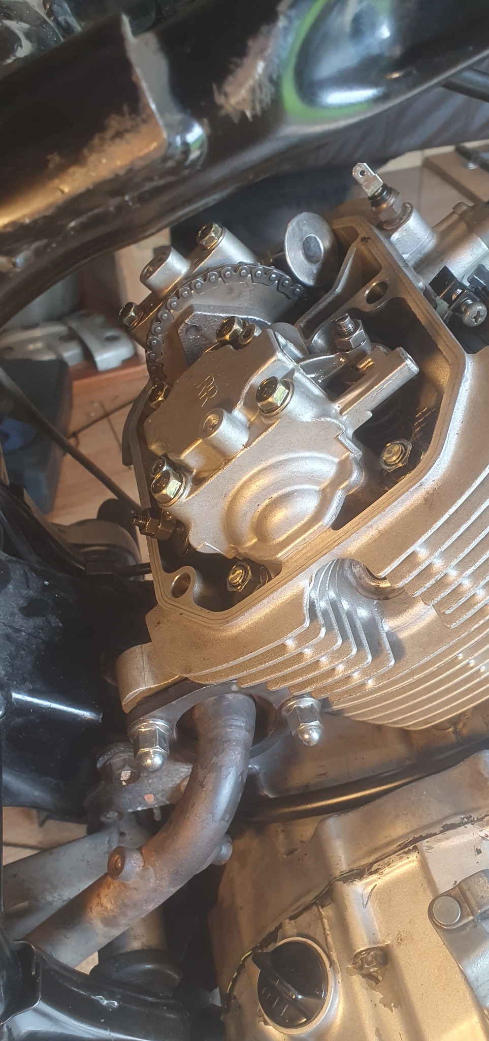 Silnik motocykla w trakcie naprawy, widoczne elementy układu rozrządu, łańcuch i pokrywa zaworów. Detaliczny widok na mechanizm z licznymi śrubami mocującymi.