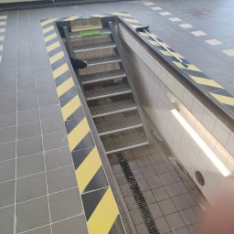 TECH-WELD - Metalowe schody kratowe prowadzące w dół, otoczone szarą i żółto-czarną płytkowaną podłogą, z widocznym odpływem liniowym i fragmentem osoby w tle.