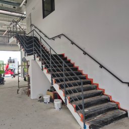 TECH-WELD - Wnętrze hali z metalowymi schodami i poręczą, zabezpieczonymi folią malarską. Widoczne wiadra z farbą i sprzęt budowlany w tle. Ujęcie z dołu, eksponujące konstrukcję.
