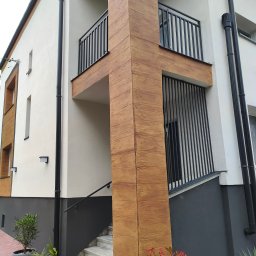 TECH-WELD - Dwupiętrowy dom z balkonami i pionowymi, czarnymi balustradami. Elewacja w kolorze złamanej bieli z elementami dekoracyjnymi imitującymi drewno. Widoczne schody wejściowe.
