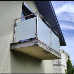 TECH-WELD - Balkon z nowoczesną balustradą ze szkła i stali nierdzewnej, zamontowaną na betonowej płycie. Widok z dołu, na tle częściowo widocznego nieba i budynku w budowie.