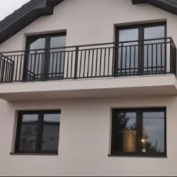 Elewacja domu z balkonem zabezpieczonym czarną, metalową konstrukcją z pionowymi szczeblami. Widoczne okna z ciemnymi ramami i grafitowy dach.
