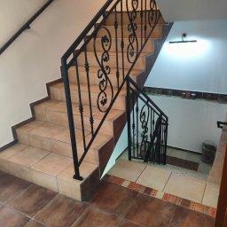 TECH-WELD - Klatka schodowa z czarną, metalową balustradą o dekoracyjnym wzorze. Schody wyłożone płytkami w odcieniach beżu i brązu. Widok z góry na dół.