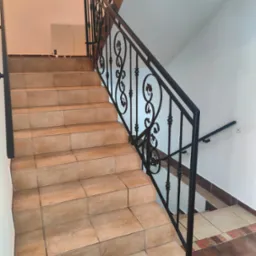 Klatka schodowa z beżowymi płytkami i czarną, zdobioną metalową poręczą. Dodatkowa poręcz na ścianie. Widok z dołu schodów, prosto na poręcz.