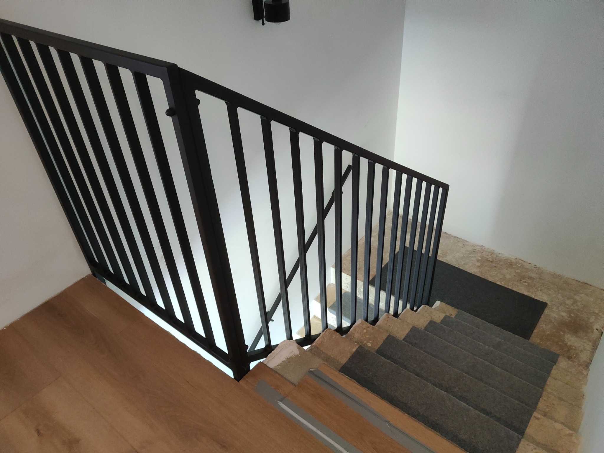 Minimalistyczna, czarna poręcz schodowa z pionowymi szczeblami, widok z góry na schody z drewnianymi i szarymi stopniami, białe ściany. Nowoczesny design.