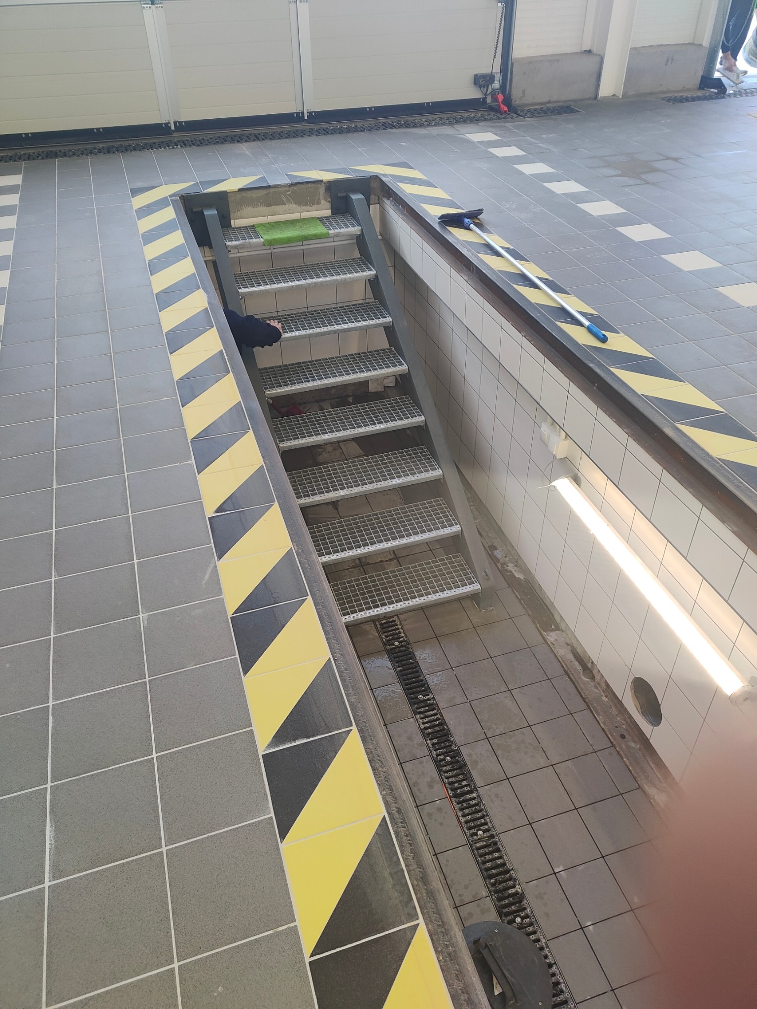 Metalowe schody kratowe prowadzące w dół, otoczone szarą i żółto-czarną płytkowaną podłogą, z widocznym odpływem liniowym i fragmentem osoby w tle.