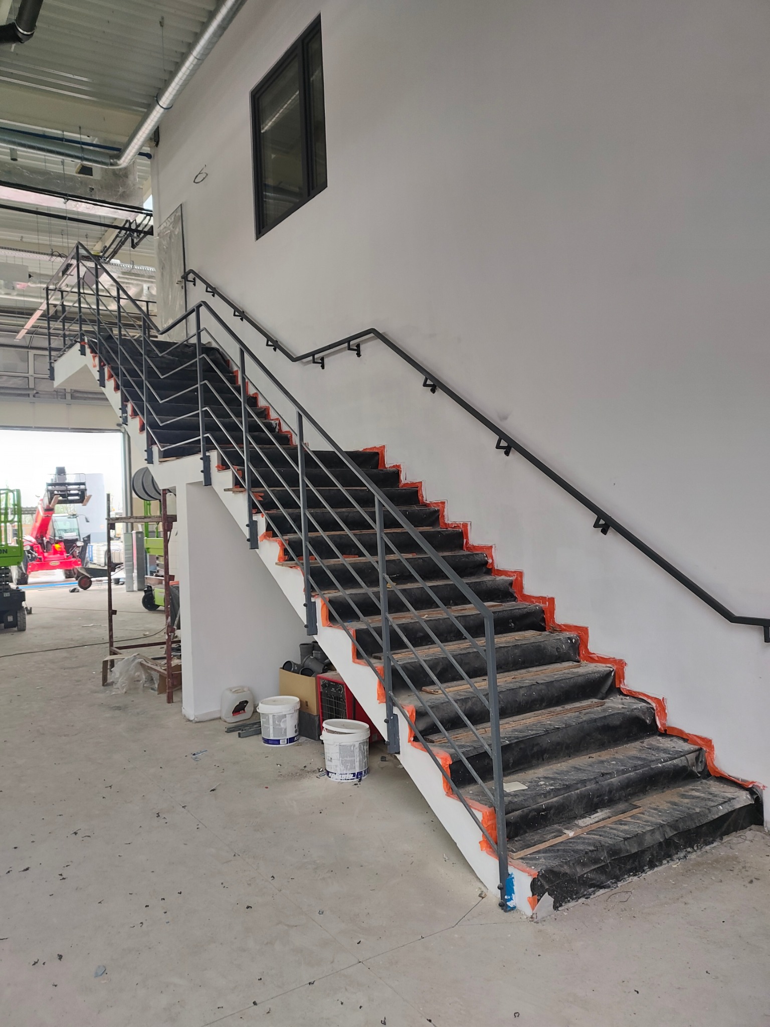 Wnętrze hali z metalowymi schodami i poręczą, zabezpieczonymi folią malarską. Widoczne wiadra z farbą i sprzęt budowlany w tle. Ujęcie z dołu, eksponujące konstrukcję.