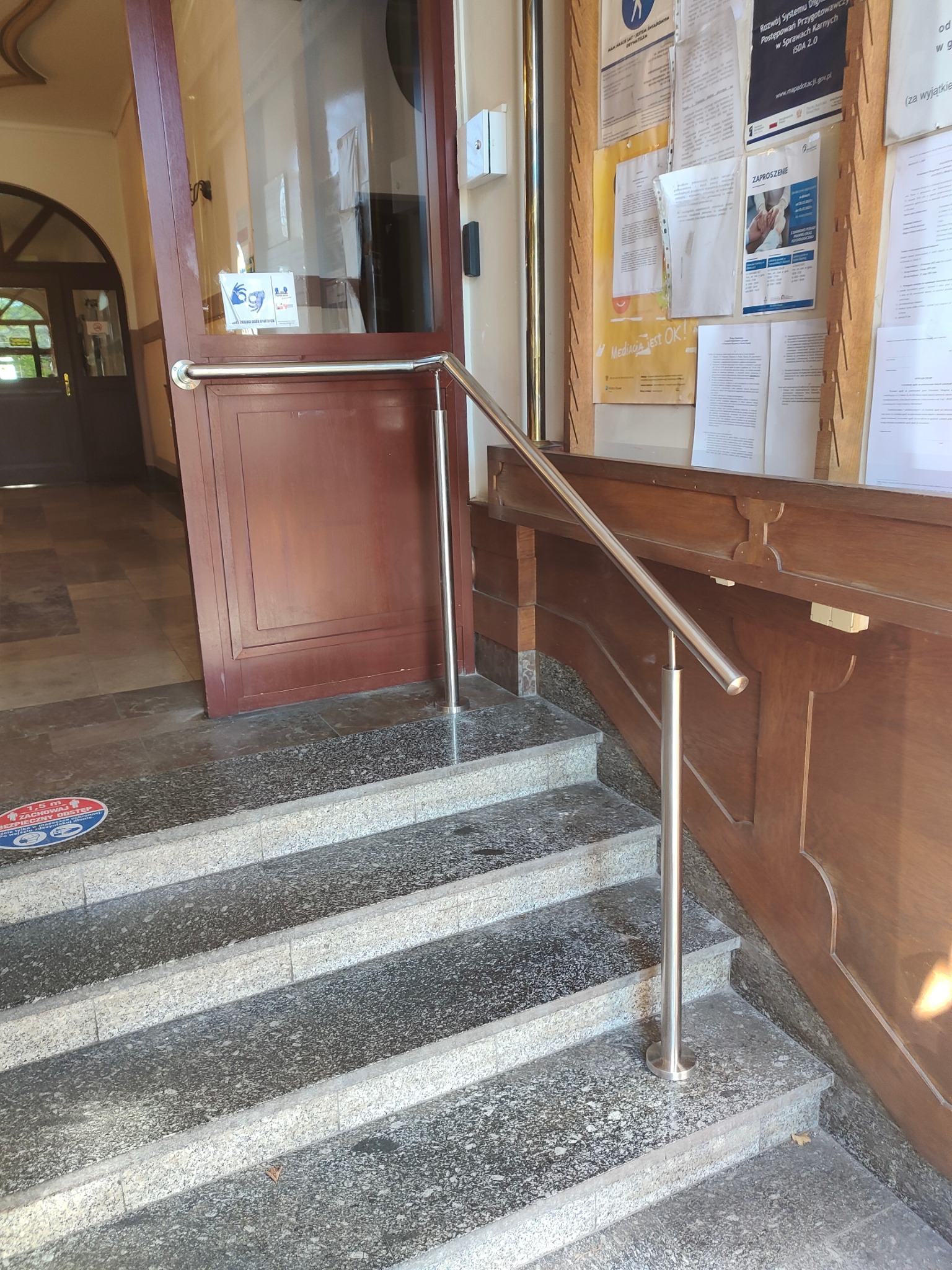 Metalowa poręcz przy schodach wejściowych do budynku, zapewniająca wsparcie. Poręcz zamontowana przy brązowych drzwiach i drewnianej boazerii. Widoczne schody z szarego lastryko.
