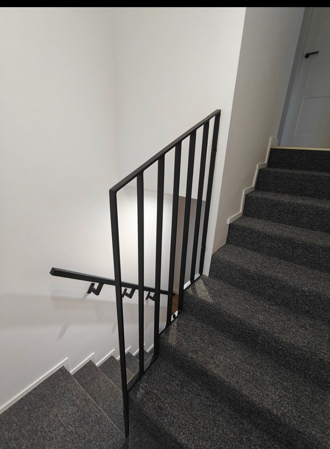 Minimalistyczna, czarna poręcz schodowa z pionowymi szczeblami, zamontowana przy białej ścianie. Widok z góry na schody pokryte szarą wykładziną.