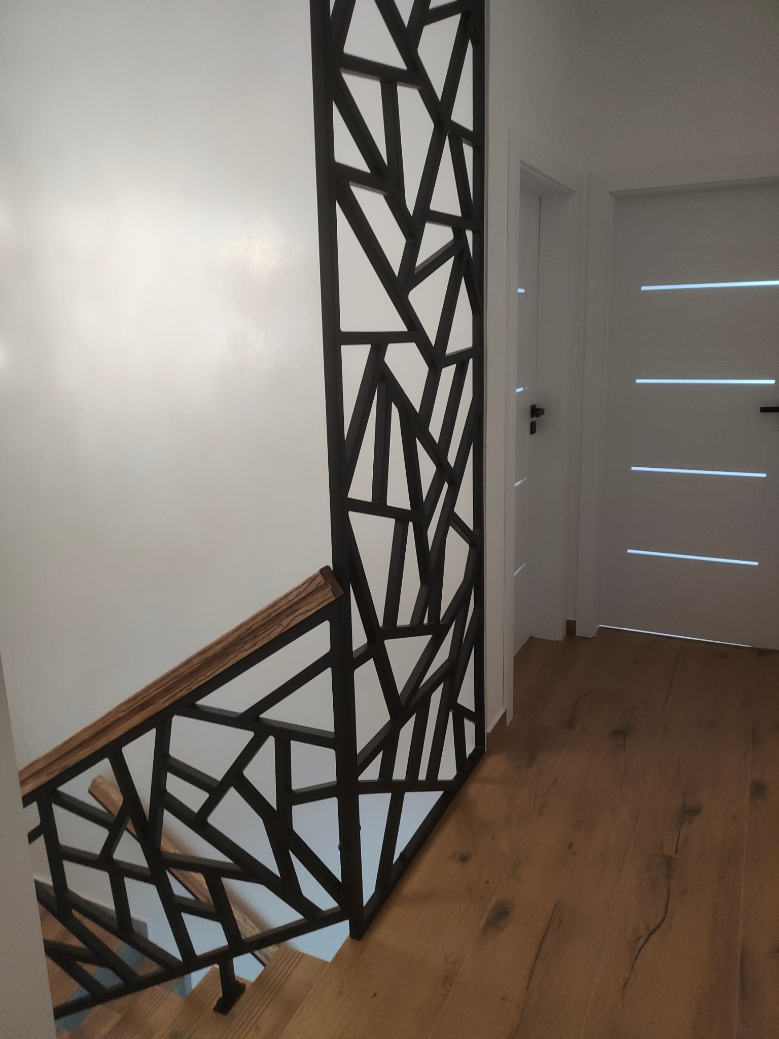 Nowoczesna balustrada metalowa z drewnianym poręczą, geometryczny wzór, jasne wnętrze z drewnianą podłogą i białymi drzwiami w tle.