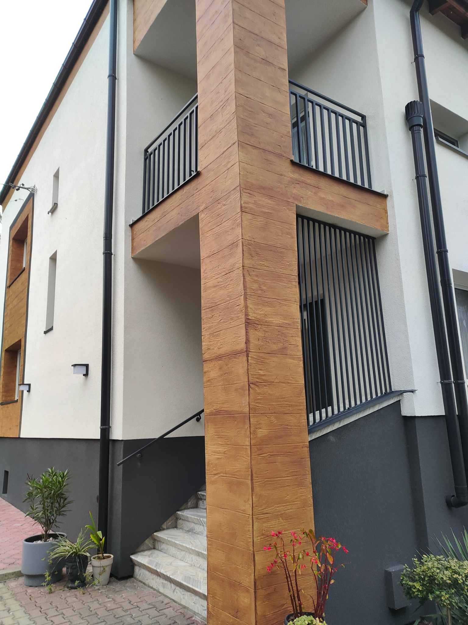 Dwupiętrowy dom z balkonami i pionowymi, czarnymi balustradami. Elewacja w kolorze złamanej bieli z elementami dekoracyjnymi imitującymi drewno. Widoczne schody wejściowe.