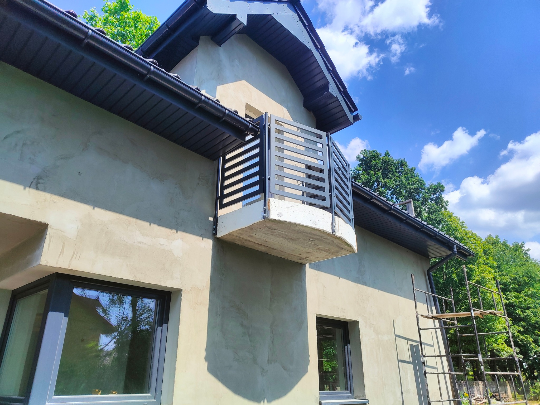 Szara, metalowa balustrada balkonowa z poziomymi szczebelkami w Będzinie. Widok z dołu na tle błękitnego nieba z chmurami i fragmentu elewacji domu.