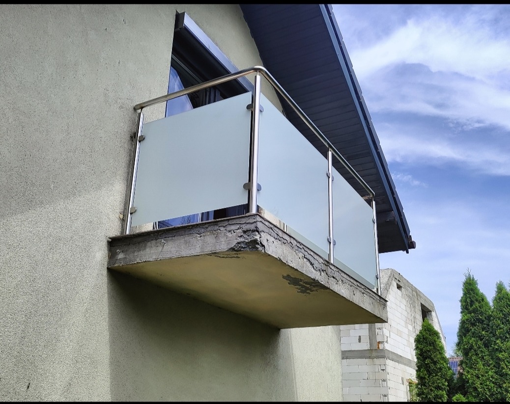 Balkon z nowoczesną balustradą ze szkła i stali nierdzewnej, zamontowaną na betonowej płycie. Widok z dołu, na tle częściowo widocznego nieba i budynku w budowie.