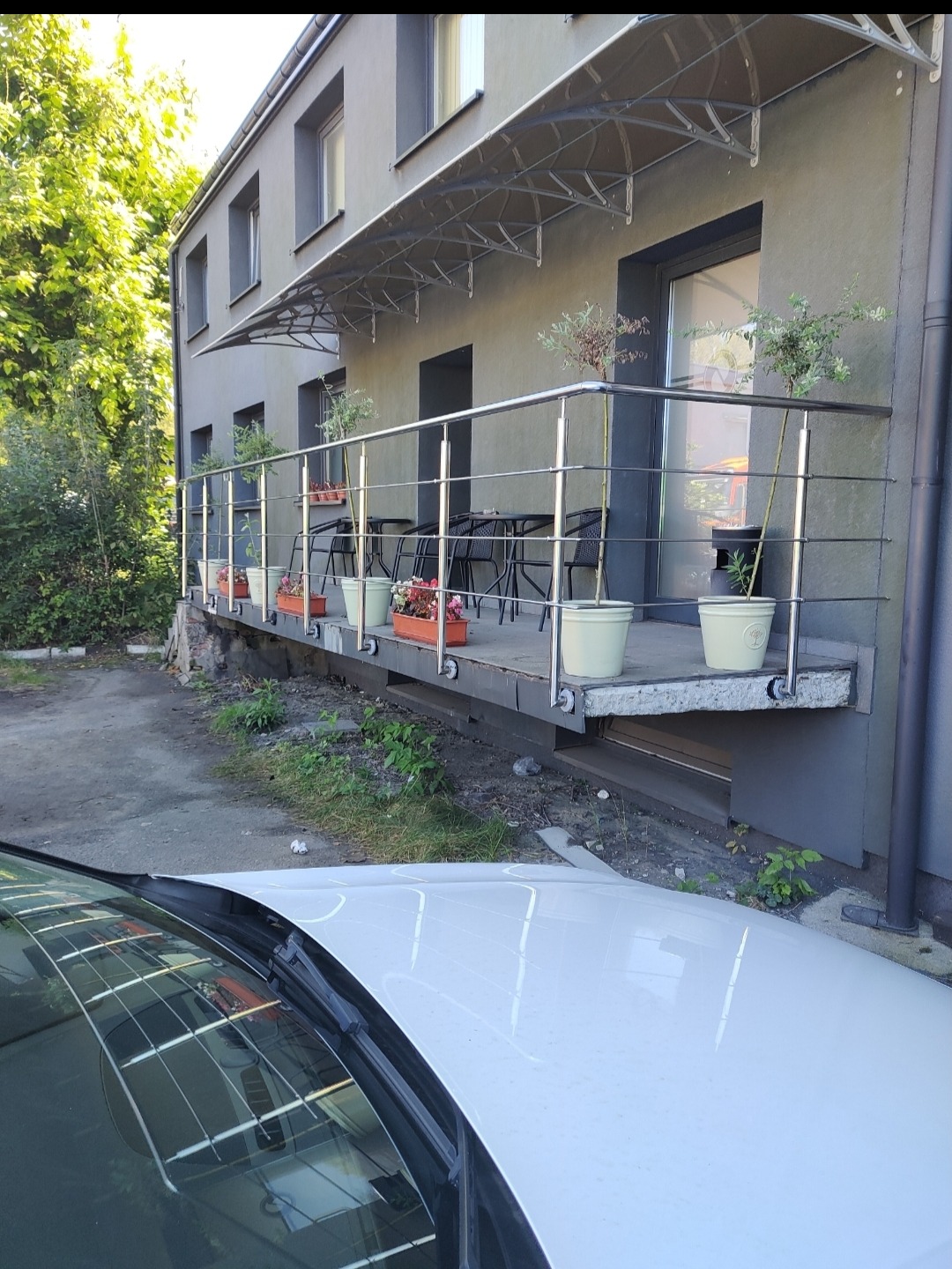 Balkon z metalową balustradą i roślinami w donicach, widok z dołu na tle szarej elewacji budynku i zieleni. Częściowo widoczny biały samochód.