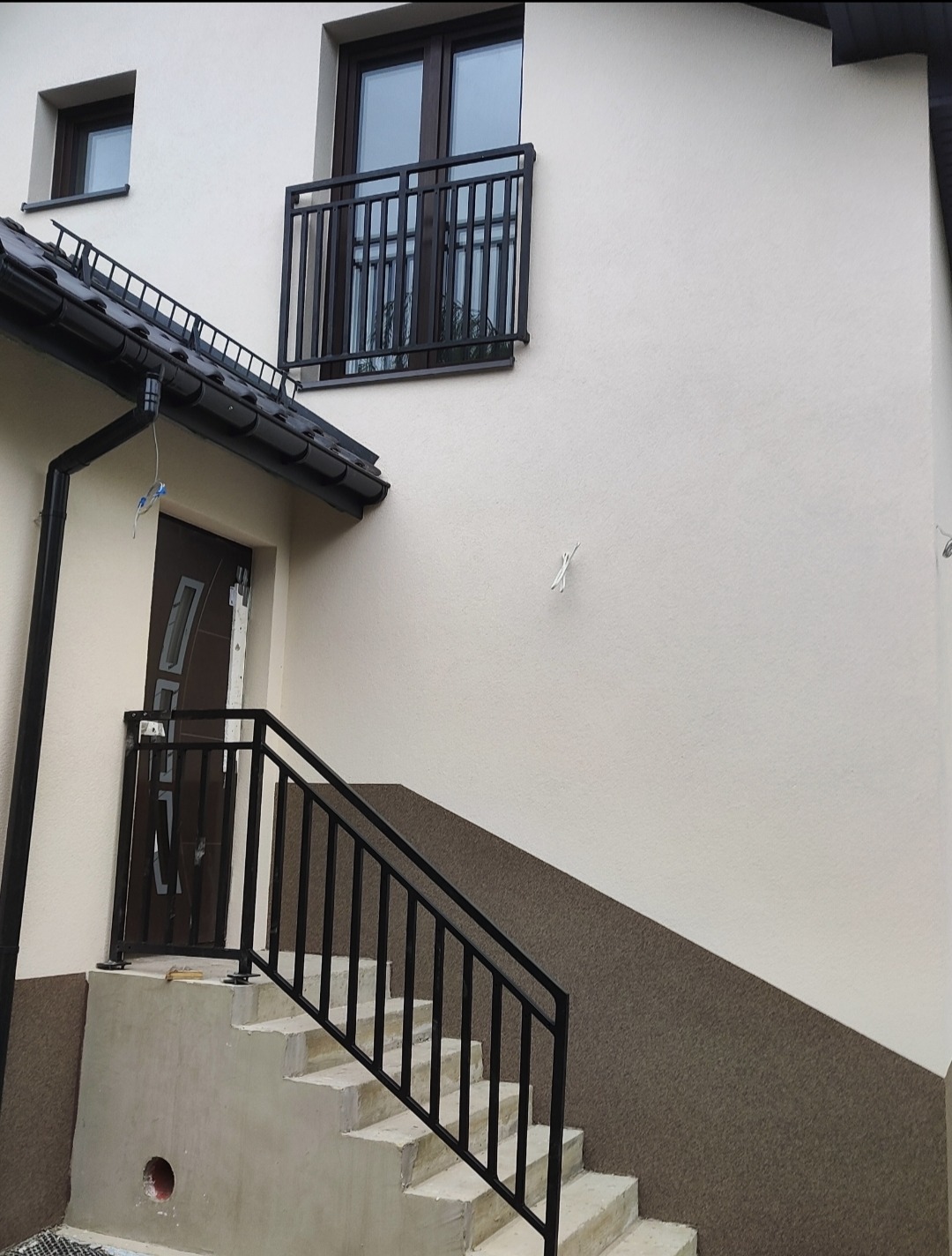 Metalowa balustrada schodowa w kolorze czarnym, prowadząca do drzwi wejściowych. Nad nimi balkon z identyczną balustradą. Elewacja domu w jasnych barwach.