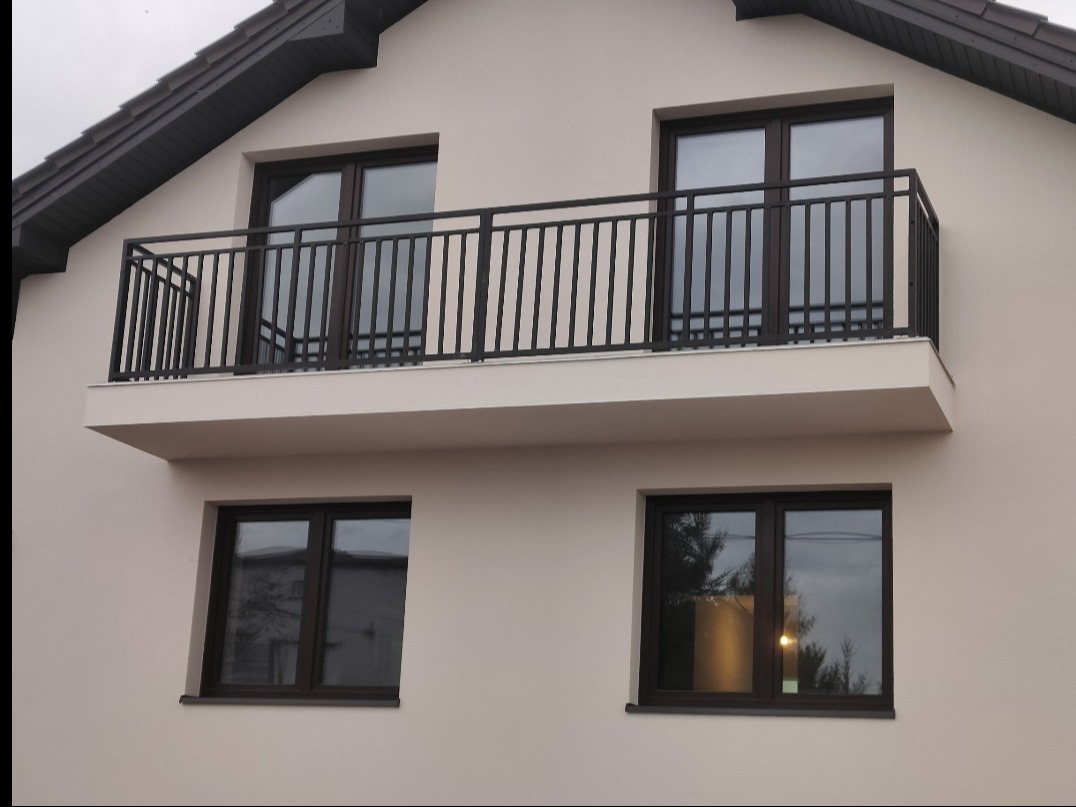Elewacja domu z balkonem zabezpieczonym czarną, metalową konstrukcją z pionowymi szczeblami. Widoczne okna z ciemnymi ramami i grafitowy dach.