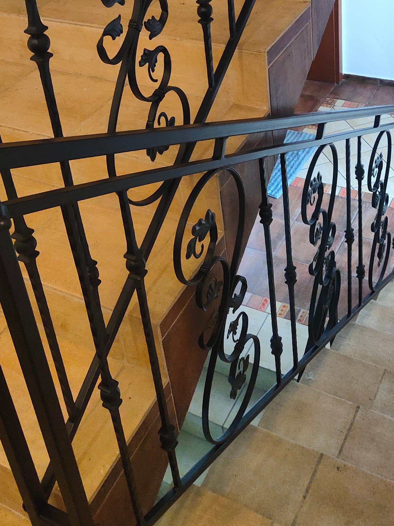 Kute, czarne poręcze schodowe z dekoracyjnymi elementami roślinnymi, widoczne z góry. Beżowe płytki schodowe w tle.