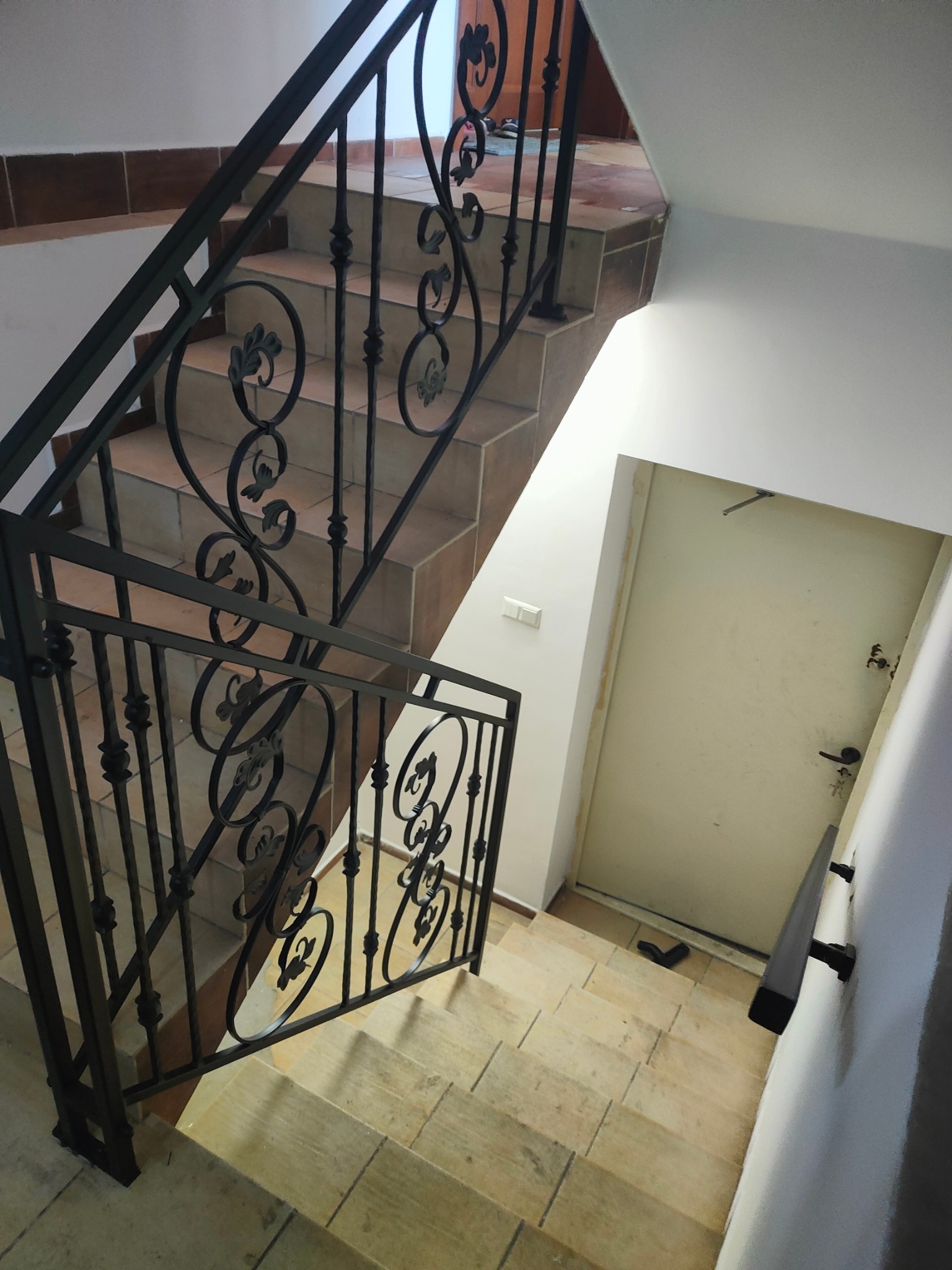 Kute, czarne balustrady schodowe z ornamentami, widok z góry. Schody wyłożone płytkami w odcieniach beżu i brązu. Białe ściany, drzwi wejściowe w tle.