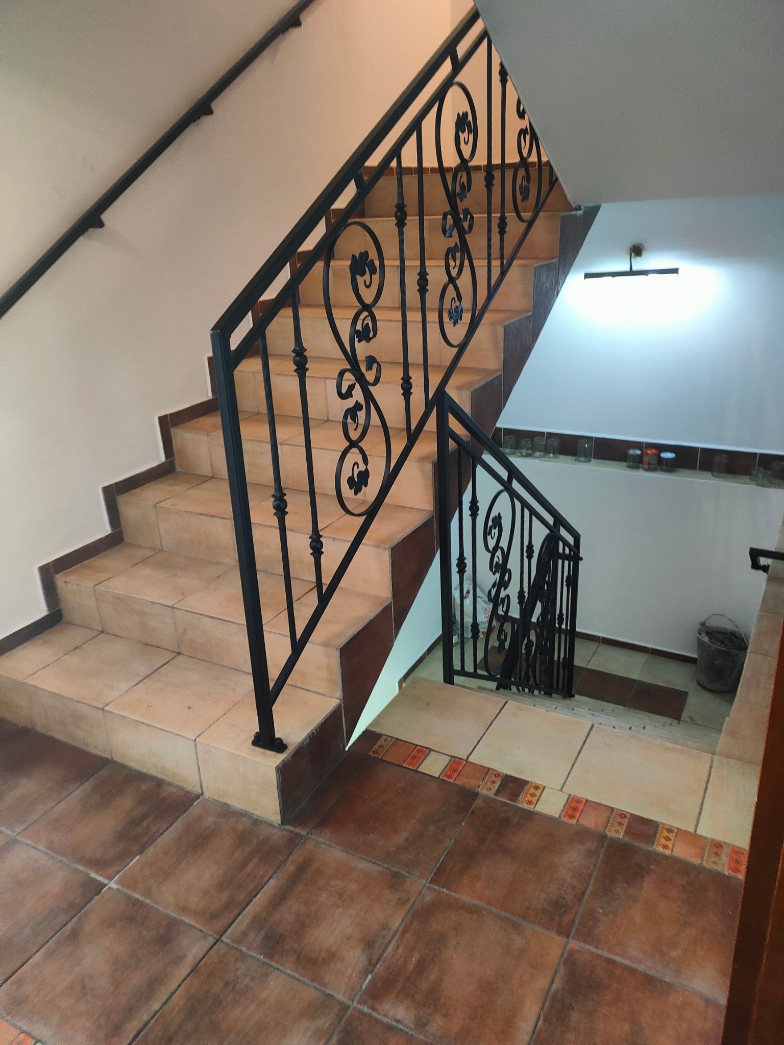 Klatka schodowa z czarną, metalową balustradą o dekoracyjnym wzorze. Schody wyłożone płytkami w odcieniach beżu i brązu. Widok z góry na dół.