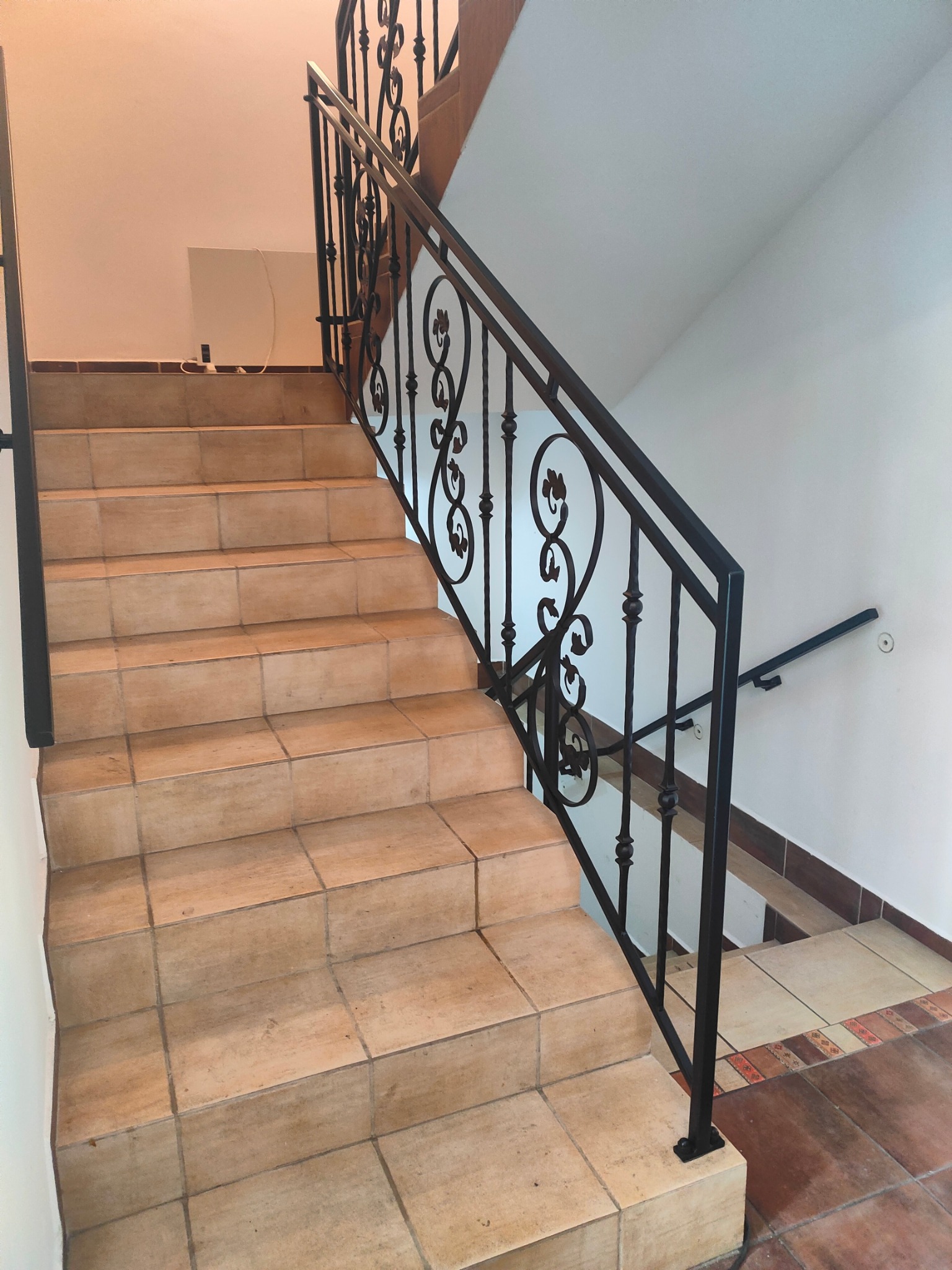 Klatka schodowa z beżowymi płytkami i czarną, zdobioną metalową poręczą. Dodatkowa poręcz na ścianie. Widok z dołu schodów, prosto na poręcz.