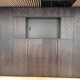 Red - Elegancka zabudowa meblowa z ciemnego drewna, wzór w jodełkę, pionowe uchwyty. Górna część z wnęką, widoczny fragment sufitu z drewnianymi listwami.