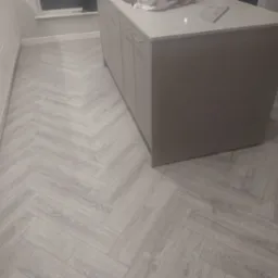 Jasnoszara podłoga ułożona w jodełkę w kuchni, widoczna wyspa kuchenna i białe szafki. Minimalistyczny design, subtelne oświetlenie.