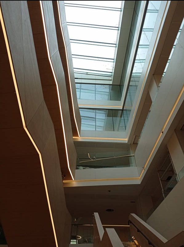 Wnętrze nowoczesnego biura z atrium. Widok z dołu na wielopoziomową przestrzeń z drewnianymi panelami i oświetleniem LED. Szklany dach wpuszcza naturalne światło.