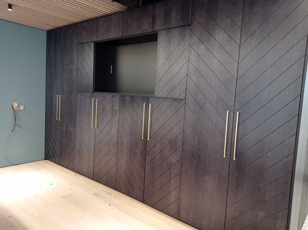 Ciemne, drewniane szafy wnękowe z chevronowym wzorem i złotymi uchwytami, w pokoju z jasną podłogą i niebieską ścianą. Widoczne gniazdko elektryczne.