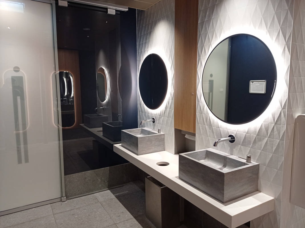 Nowoczesna łazienka publiczna z umywalkami w kształcie prostopadłościanów i okrągłymi lustrami z podświetleniem LED. Białe ściany z trójkątną fakturą. Minimalistyczny design.