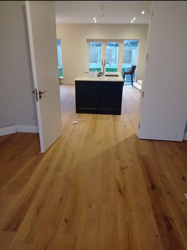 Jasna, drewniana podłoga z widoczną wyspą kuchenną i otwartymi drzwiami. Nowe panele podłogowe w jasnym odcieniu drewna, świeżo po ułożeniu w przestronnym wnętrzu.