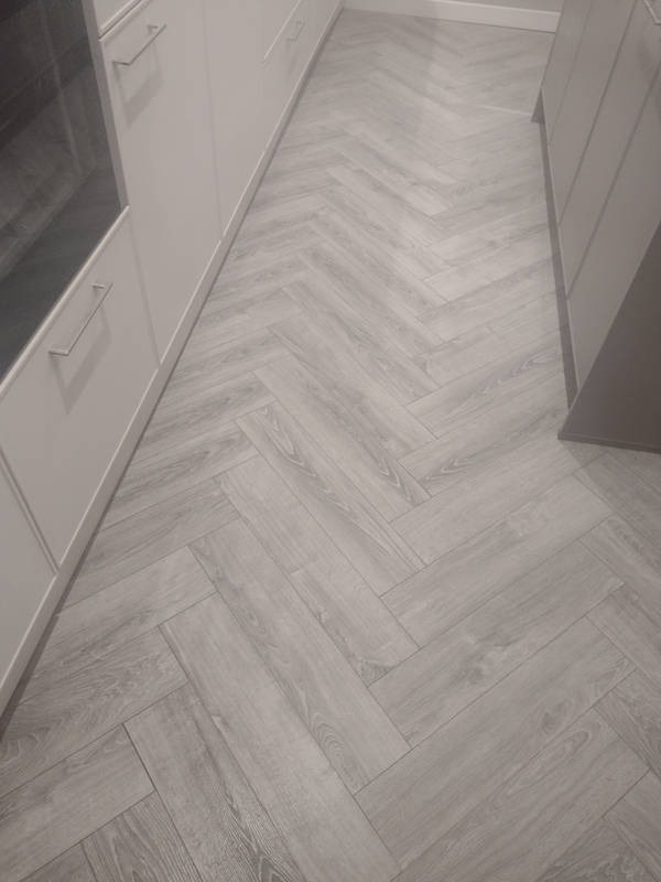 Jasnoszara podłoga z paneli ułożonych we wzór jodełki, widoczna przy białych szafkach kuchennych z metalowymi uchwytami. Minimalistyczny wystrój.